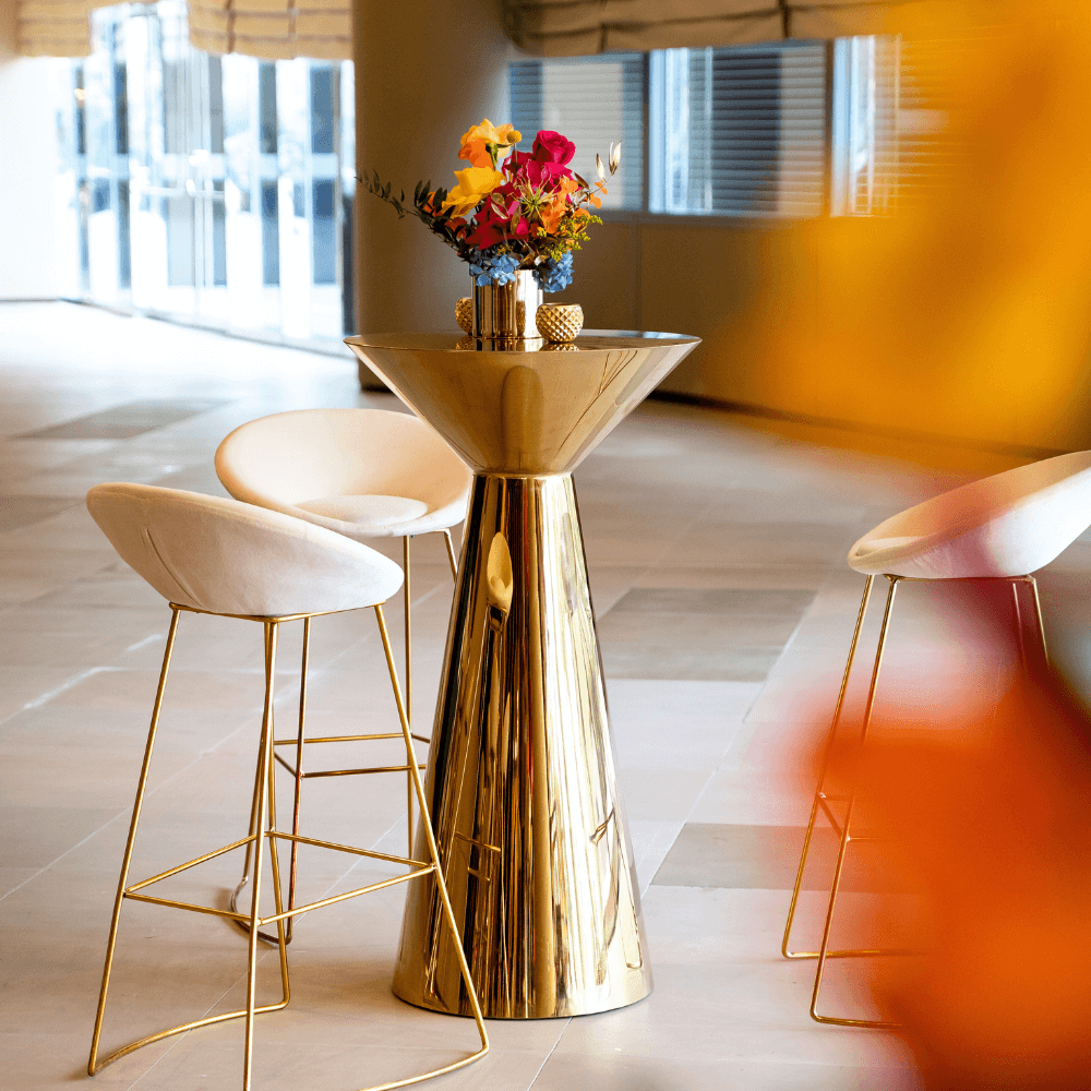 Golden Glam- Cocktail Setup featuring a table with a flower centerpiece and eight gold bar stools with white cushions, ideal for hosting events.