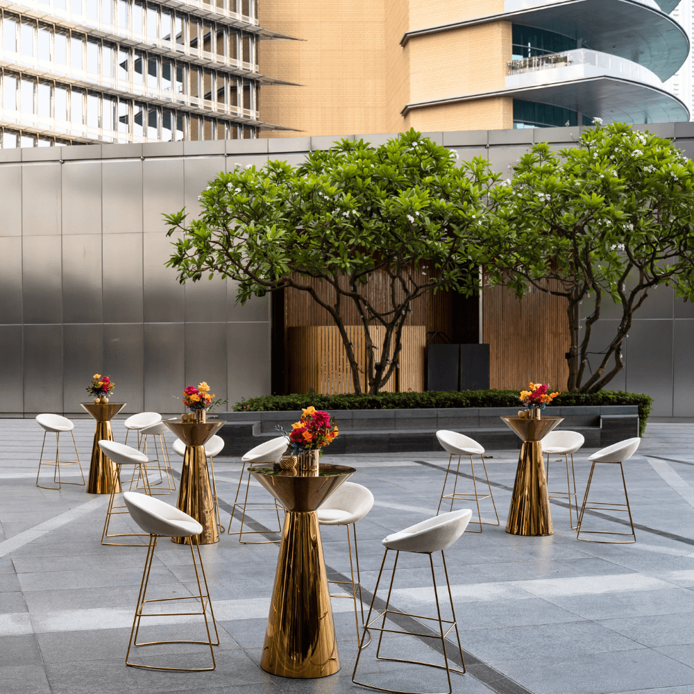 Golden Glam-Cocktail Setup featuring a table with eight white chairs with gold legs and a flower centerpiece in an elegant courtyard setting.