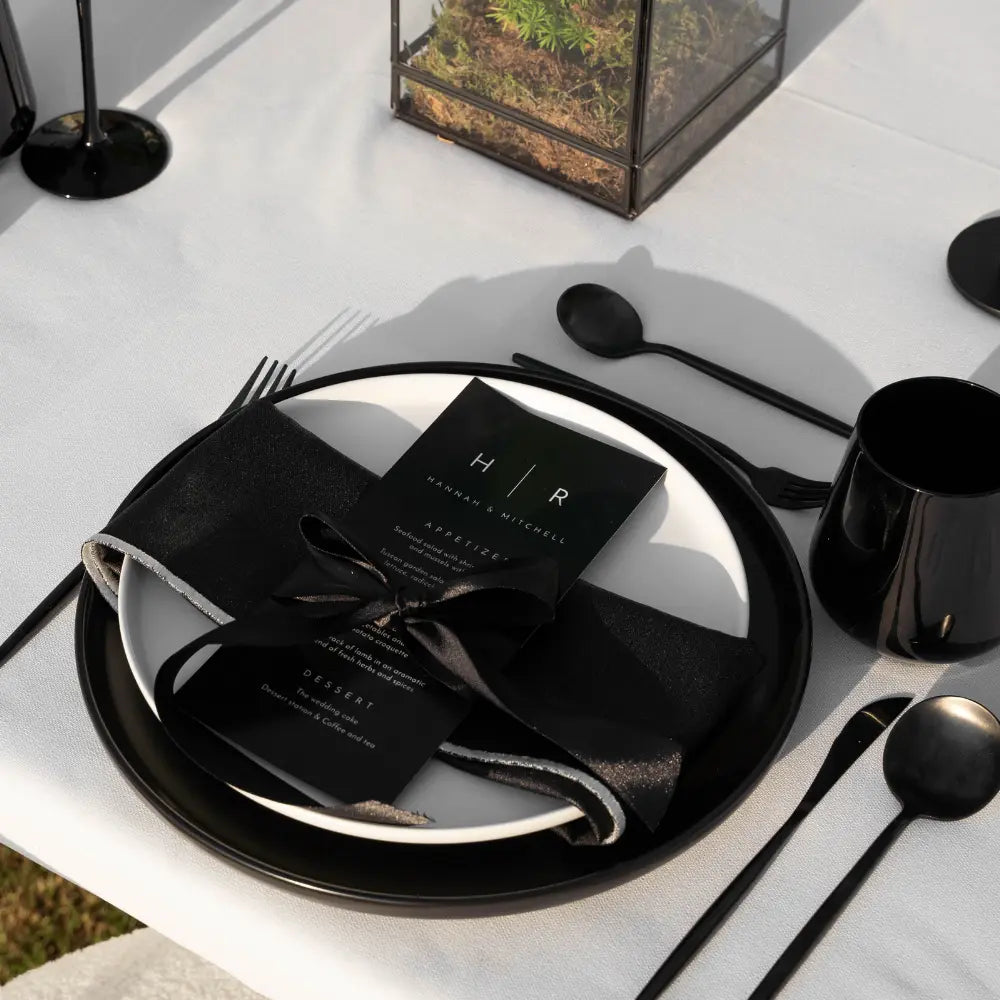 CERAMIC PLATES - BLACK, elegantly stacked with a black charger and white dinner plate, accented by matte-black flatware and a centerpiece terrarium, perfect for sophisticated events.