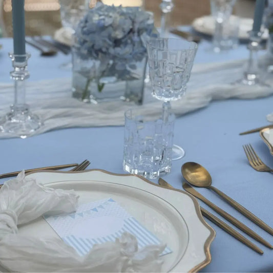 White Lotus Collection charger plate with gold trim, paired with elegant gold flatware and crystal glasses, set on a soft blue tablecloth for upscale dining.