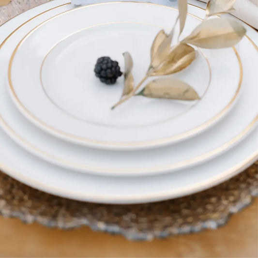 MODERN CHINA W/ GOLD RIM PLATES stacked neatly with a gold-painted sprig and blackberry on top, resting on a textured gold charger, ideal for premium event settings.