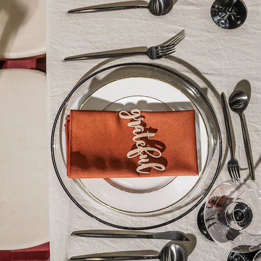 Festive place setting featuring the Clear Plate W/Black Rim Collection, with a grateful place card, burnt-orange napkin, and stainless-steel flatware on a cream tablecloth.