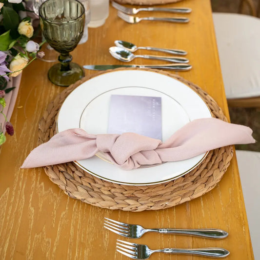 CANE PLACEMAT: A woven rattan charger beneath a white plate with a pink napkin, stainless-steel flatware, and a green glass goblet on a wooden table.