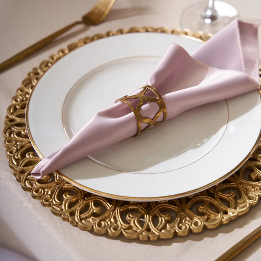 CHARGER PLATE - BRASS: An ornate gold charger plate beneath a white porcelain dinner plate, enhancing an elegant table setting with a pink napkin and gold-toned fork.
