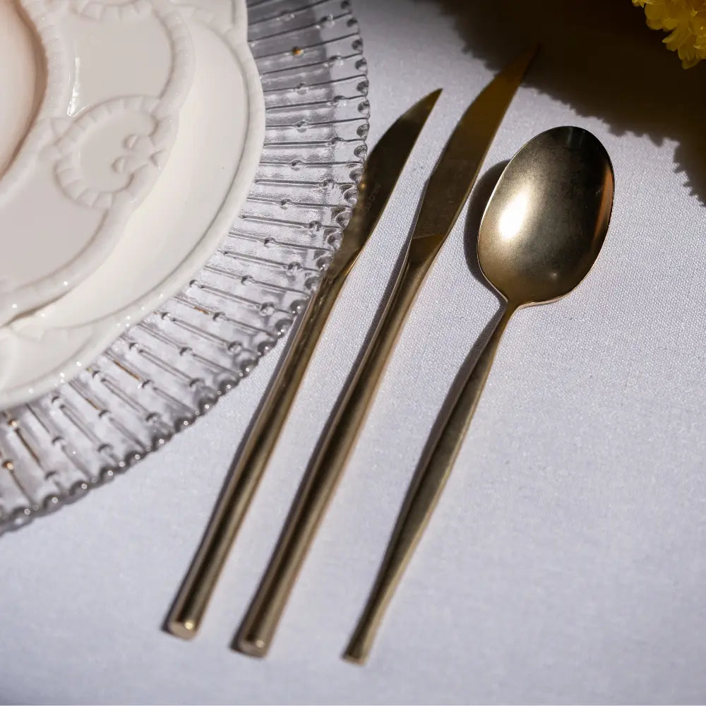 CUTLERY MODERN CHAMPAGNE: Gold-tone spoon and knives elegantly arranged beside embossed china plate on glass charger, enhancing any dinner setting.