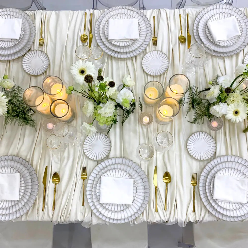 SCALLOPED LIKE PLATES elegantly layered on a formal banquet table with gold-tone flatware, clear glasses, ivory candles, and white-green floral arrangements.