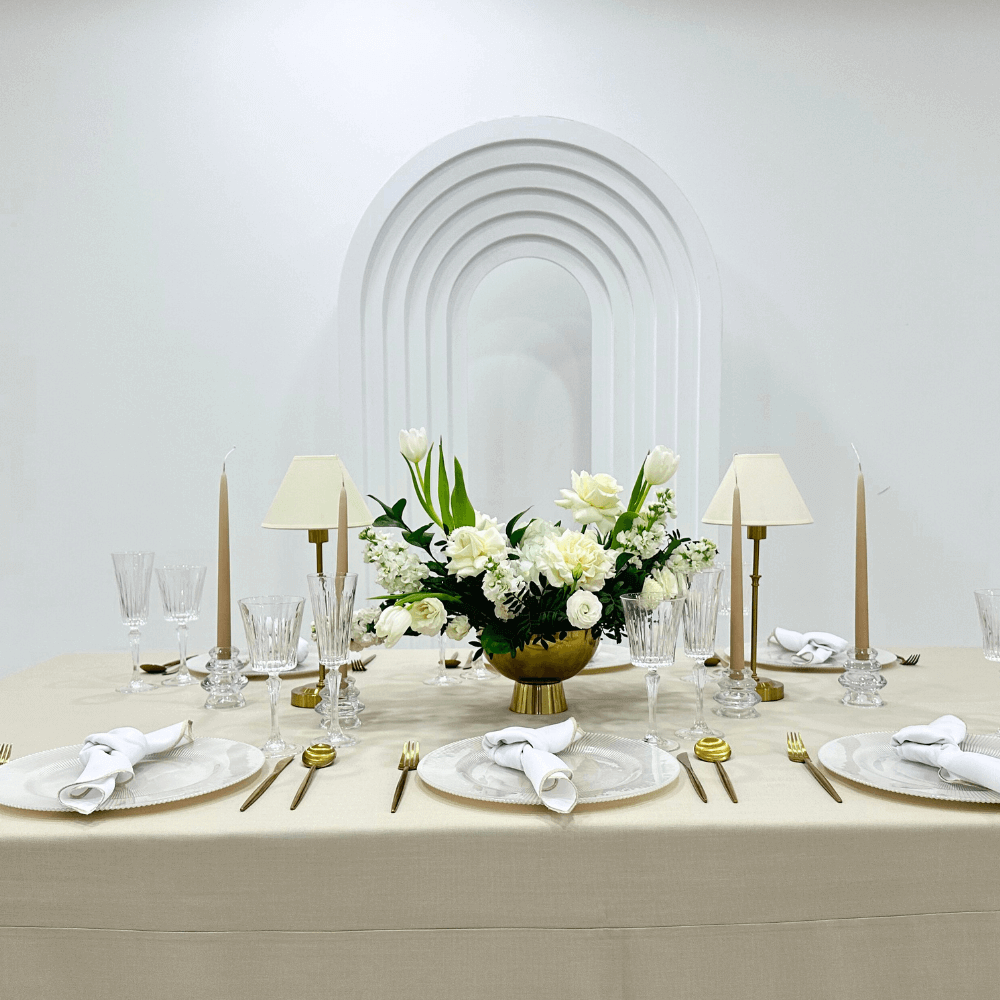 Sand Elegance- Dining Setup: table adorned with a flower centerpiece, candles, gold cutlery, charger plates, and wine glasses for an elegant event setting.