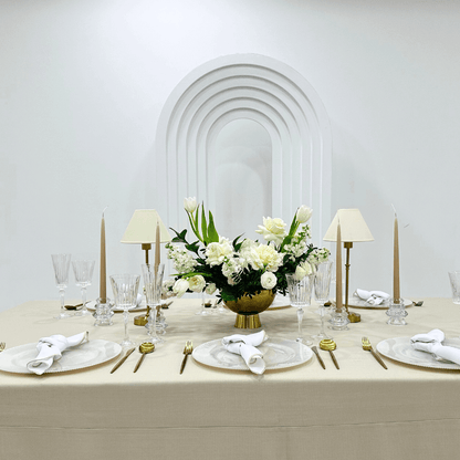 Sand Elegance- Dining Setup: table adorned with a flower centerpiece, candles, gold cutlery, charger plates, and wine glasses for an elegant event setting.