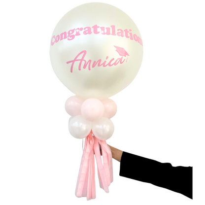 Personalized Balloon Wand featuring a large pearly-white balloon with Congratulations Annica and a mortarboard, surrounded by smaller balloons and pink tassels, held by a hand.