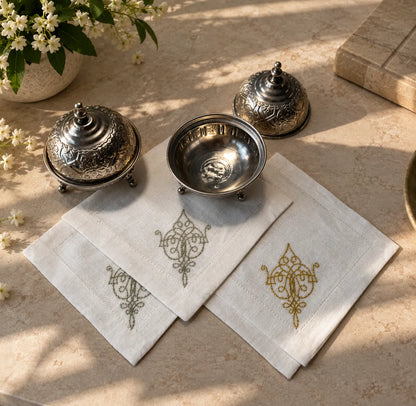 Three white embroidered cotton napkins with ornate gold and gray motifs, arranged on a beige stone surface alongside decorative silver metal dishes and small white flowers.