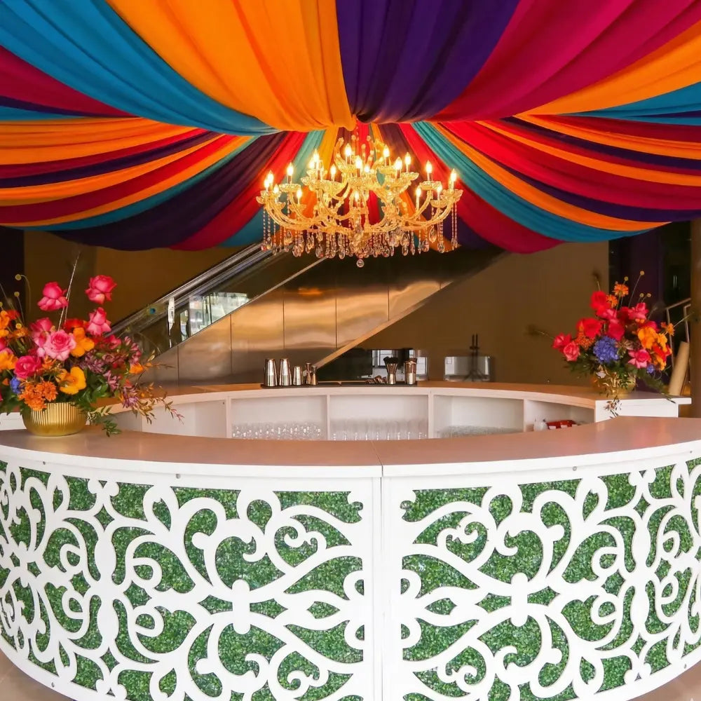 Arabesque Bar Unit with white scrollwork panels and green foliage, adorned with vibrant overhead drapery and floral arrangements, ideal for enhancing event spaces.