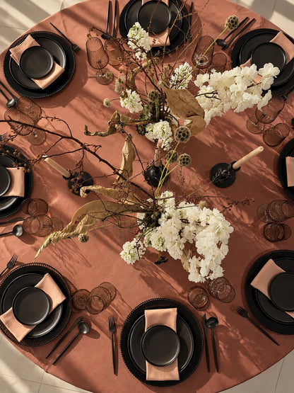 Overhead view of a styled dining table set with black plates, blush pink napkins, clear textured glassware, and a rustic floral centerpiece with white blossoms and dried botanicals on a rust-colored tablecloth.