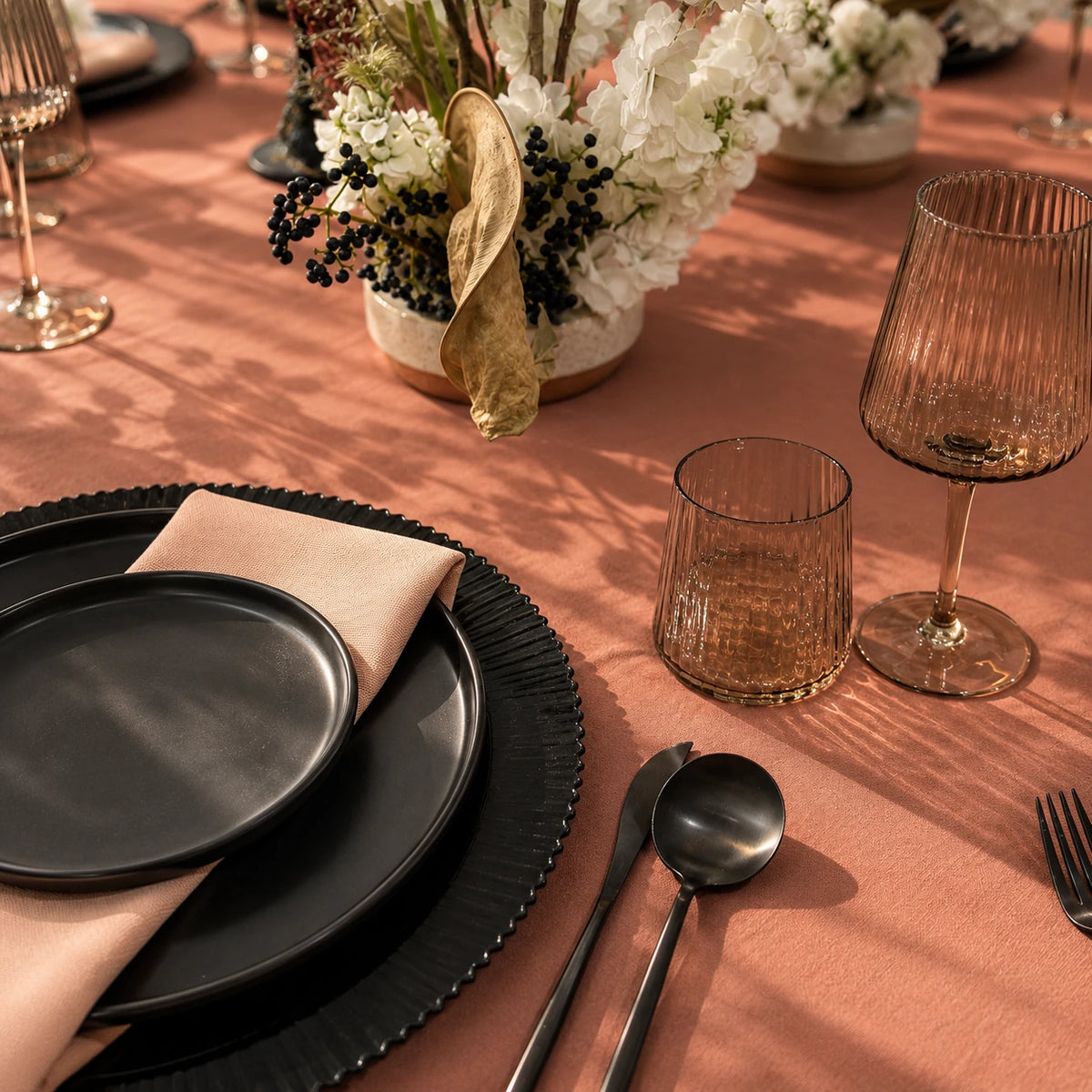 A styled table setting with matte black plates, a blush pink napkin, clear ribbed glass tumblers, and silver flatware on a dusty rose tablecloth, with a floral centerpiece in the background.