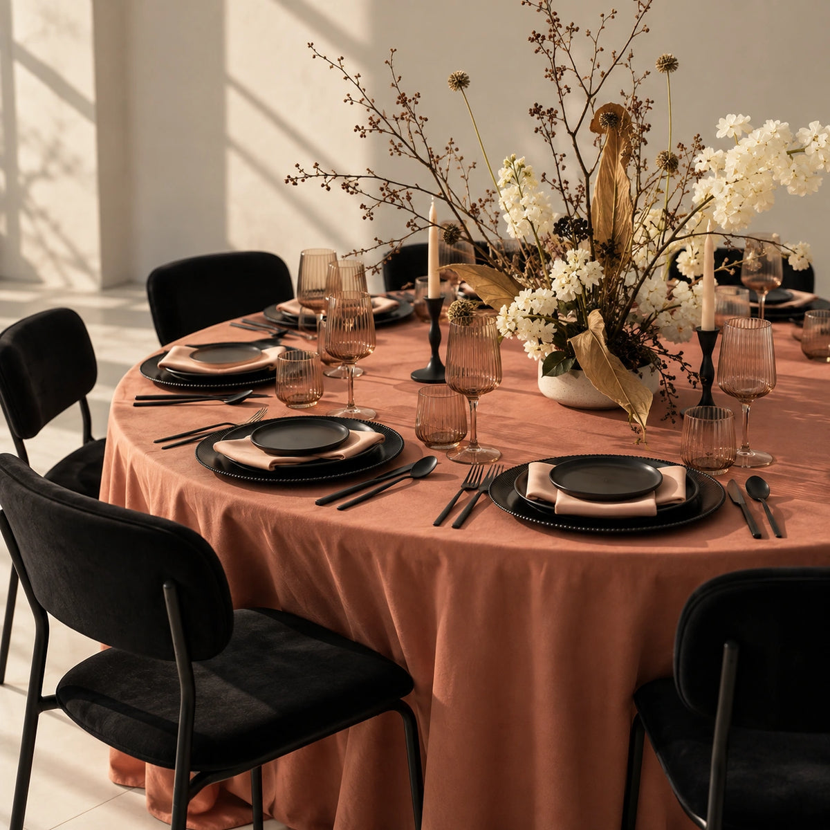 A styled round dining table set for six with a peach tablecloth, black plates, black chairs, clear glassware, and a central floral arrangement with white flowers and dried botanicals.