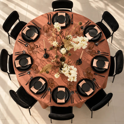 Overhead view of a round dining table set for eight, featuring a terracotta-colored tablecloth, black plates, black napkins, black glassware, and a central floral arrangement on a light tiled floor.