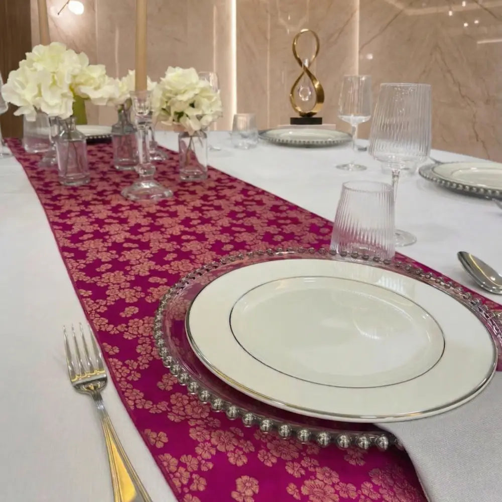 Brocade Table Runners elegantly adorn a dining table, featuring a fuchsia-pink runner with gold floral motifs, complemented by refined dinnerware and white floral arrangements.