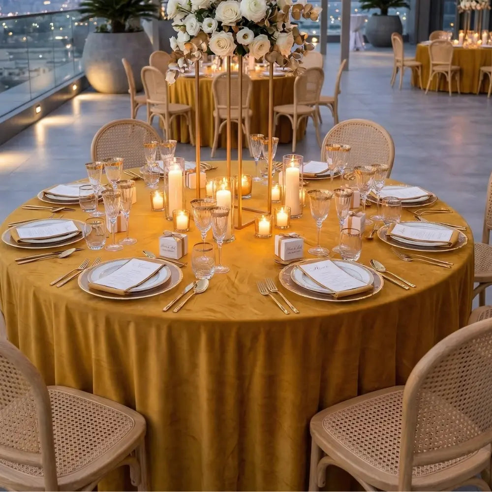 Decorated round table with yellow tablecloth, candles, and place settings in an outdoor setting.