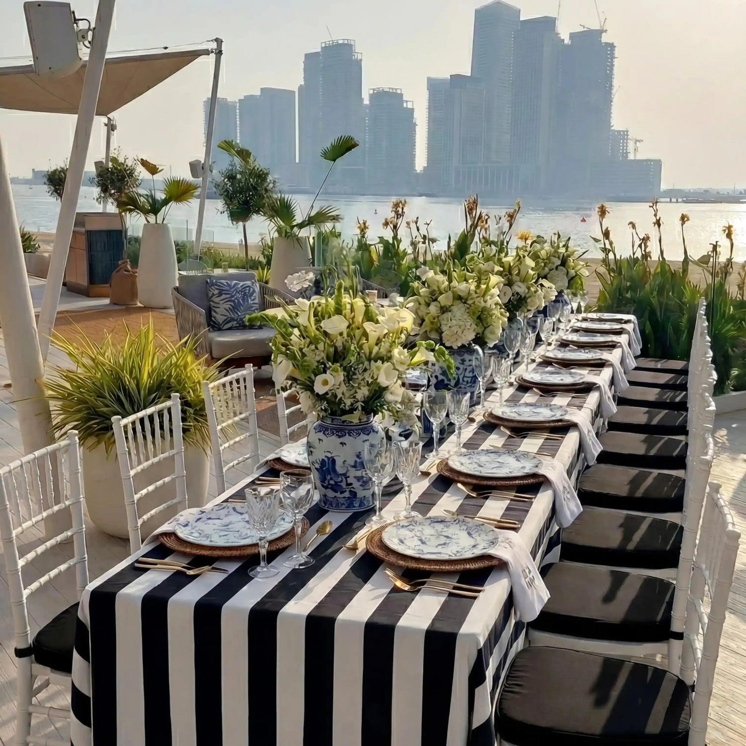 Monochrome Linen Tablecloth elegantly draped on a seaside event table with black-and-white stripes, enhancing the sophisticated al fresco dining setup amidst floral arrangements and city skyline backdrop.