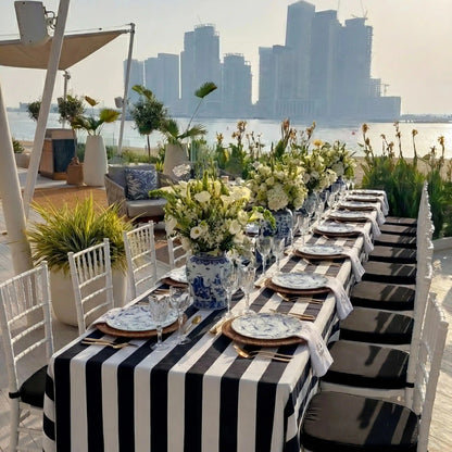 Monochrome Linen Tablecloth elegantly draped on a seaside event table with black-and-white stripes, enhancing the sophisticated al fresco dining setup amidst floral arrangements and city skyline backdrop.