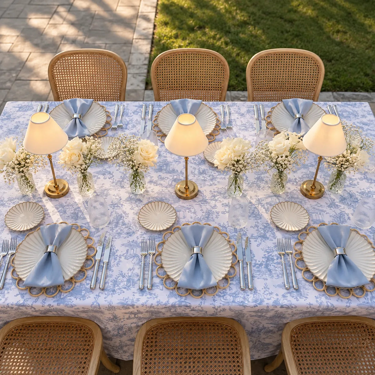 An outdoor dining table set for six with a blue and white toile patterned tablecloth, matching napkins, scalloped plates, gold-toned cutlery, small table lamps, and floral centerpieces. Woven cane chairs are arranged around the table, with a paved patio and green lawn in the background.