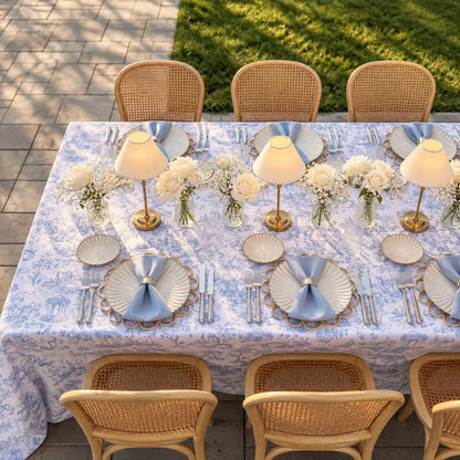 An outdoor dining table set with a light blue and white toile patterned tablecloth, matching plates, folded napkins, silverware, small table lamps, and floral centerpieces, surrounded by woven rattan chairs on a paved patio with grass in the background.