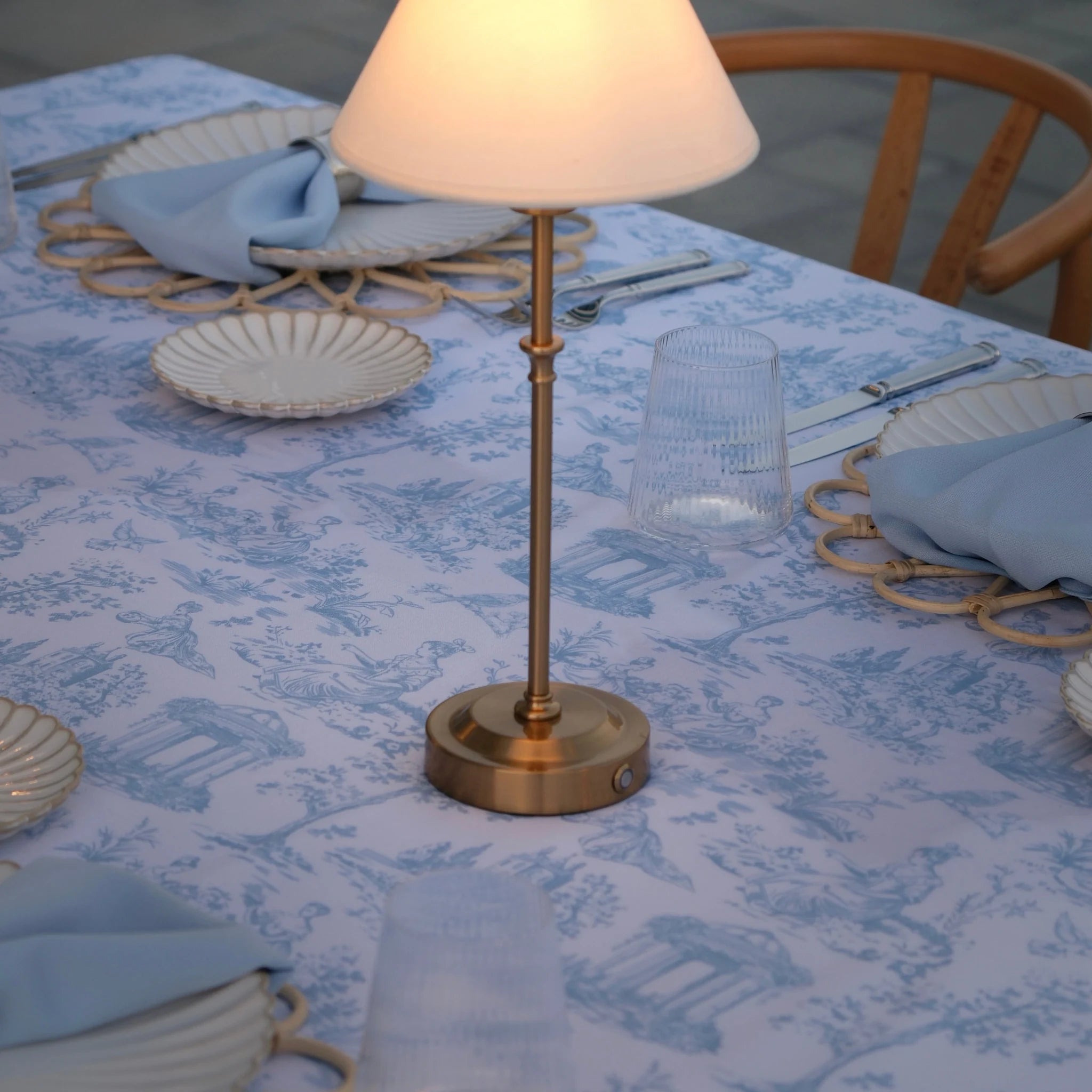 A blue-and-white toile patterned tablecloth, surrounded by plates, folded blue napkins, and a glass in an outdoor dining setting.