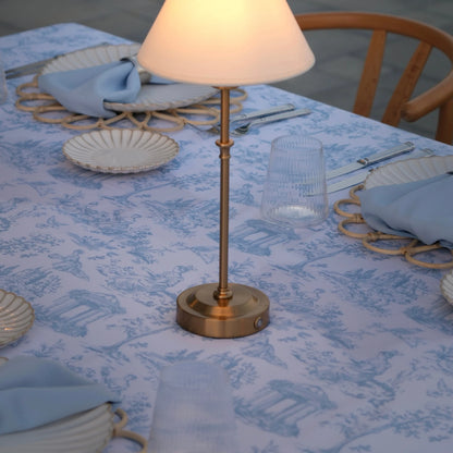 A blue-and-white toile patterned tablecloth, surrounded by plates, folded blue napkins, and a glass in an outdoor dining setting.