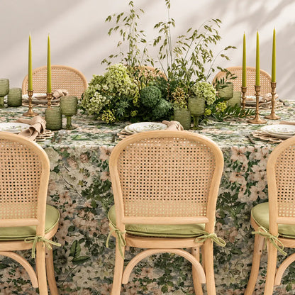 A styled dining table setup with a green and beige floral tablecloth, woven cane chairs with sage green cushions, green glassware, tall green taper candles, and lush floral centerpieces.