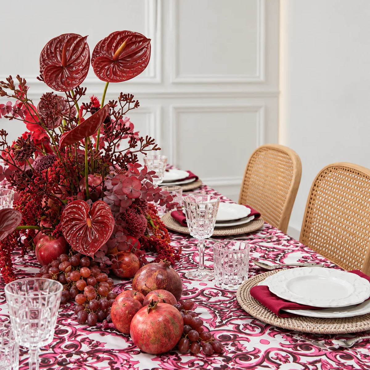 A styled dining table set with a bold magenta and white patterned tablecloth, woven placemats, white plates, clear cut glassware, red napkins, and a large red floral centerpiece with pomegranates in a bright room.