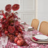 A styled dining table set with a bold magenta and white patterned tablecloth, woven placemats, white plates, clear cut glassware, red napkins, and a large red floral centerpiece with pomegranates in a bright room.