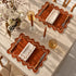 Two scalloped rust-orange placemats on a cream tablecloth, styled with white napkins, gold-toned glassware, wooden utensils, and a floral centerpiece in a bright dining setting.
