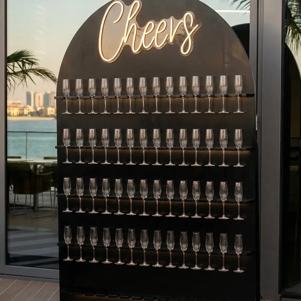 Freestanding CHAMPAGNE WALL with arch top, five shelves holding champagne flutes, and a Cheers sign above, ideal for events.