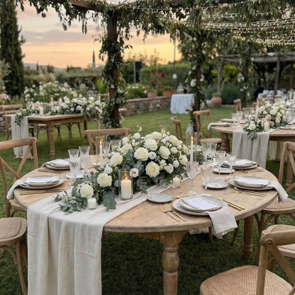 Classic Wood Round Dining Table set for an outdoor event under a pergola. Adorned with a neutral linen runner, floral centerpiece, and gold flatware.