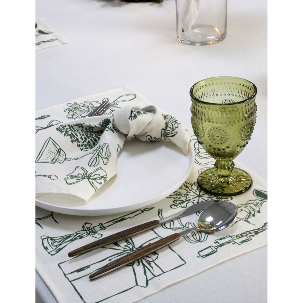 Holiday Icon Linen Placemat- 4 per pack, featuring festive green motifs, underpins a white dinner plate, enhancing holiday elegance with matching decor and wooden utensils.