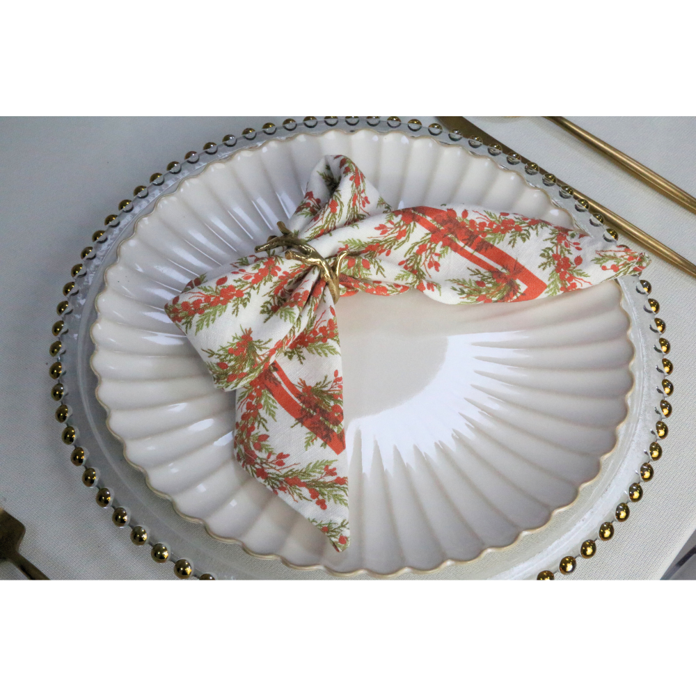 Holly &amp; Pine Linen Napkin, featuring red and green botanical motifs, elegantly folded with gold twig-style napkin rings, enhances festive table settings with luxury and warmth.
