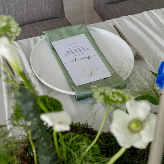 Classic Baby Shower Dinner Setup with a sage-green napkin, printed card on a white plate, surrounded by elegant floral arrangements and cutlery, exemplifying modern elegance.