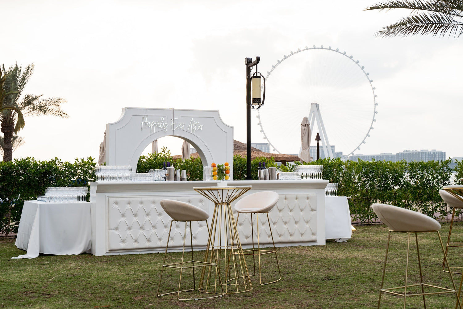 Cream and gold base bar stool with gold legs, ideal for events, weddings, and corporate functions, featured near a white bar with a Ferris wheel backdrop.