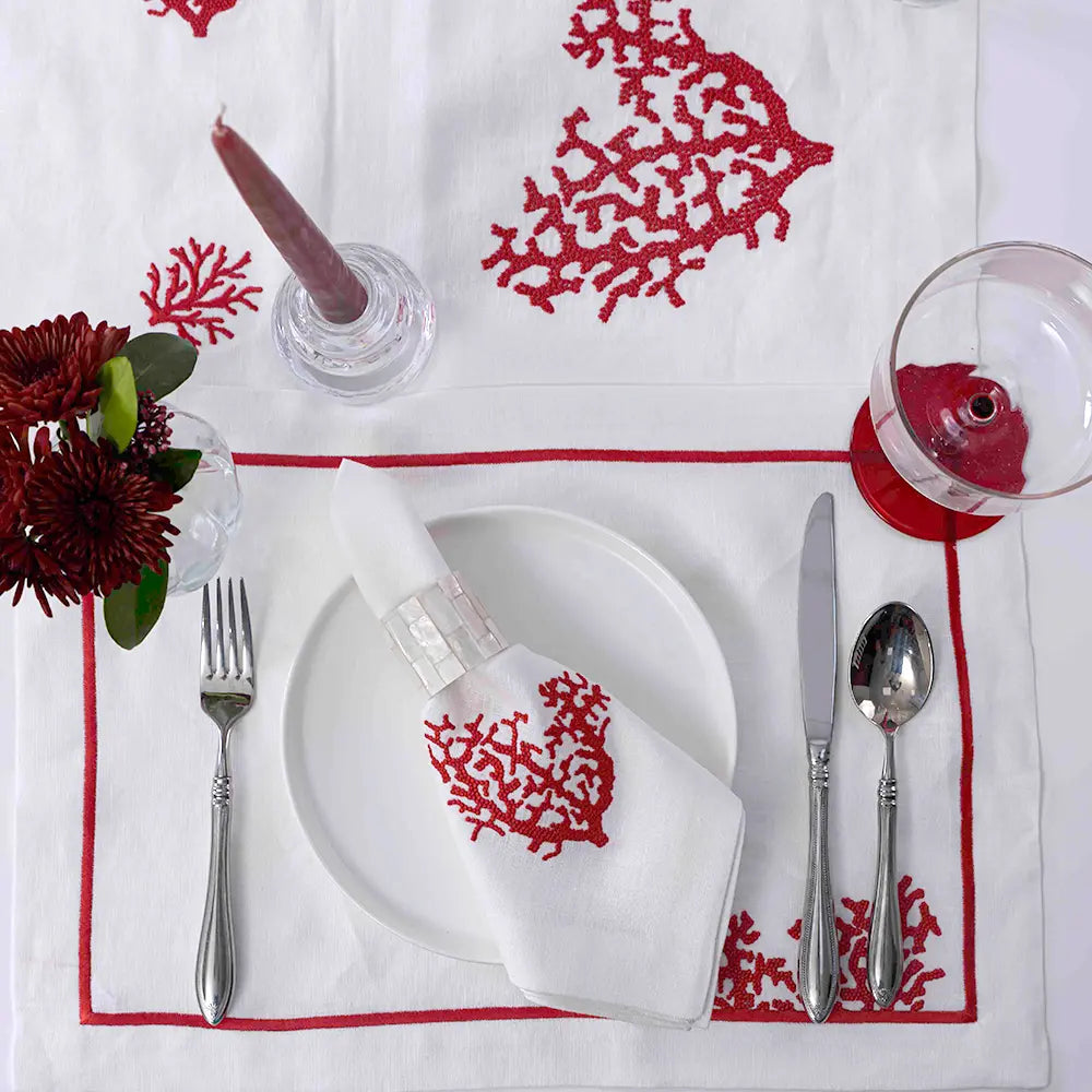 Dining table setting with red coral patterns on a white tablecloth, including cutlery and a candle.