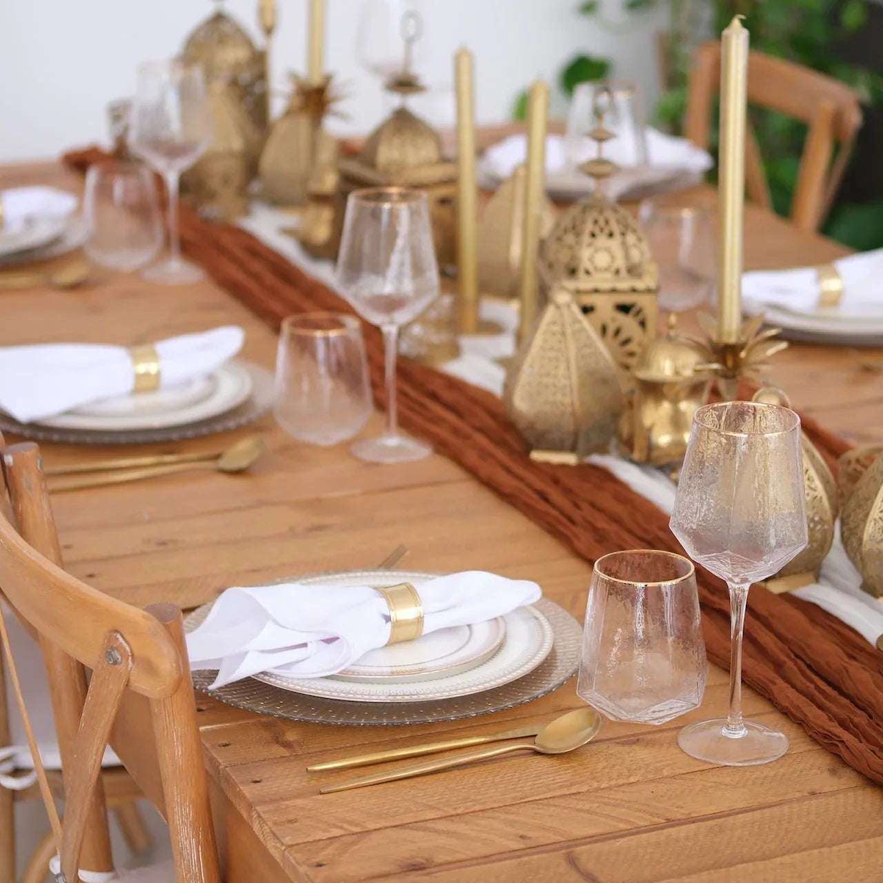 The Noor Royale - Dinner Setup: A wooden dining table elegantly set for eight, featuring gold cutlery, ornate lanterns, and crossback chairs, ideal for sophisticated gatherings.