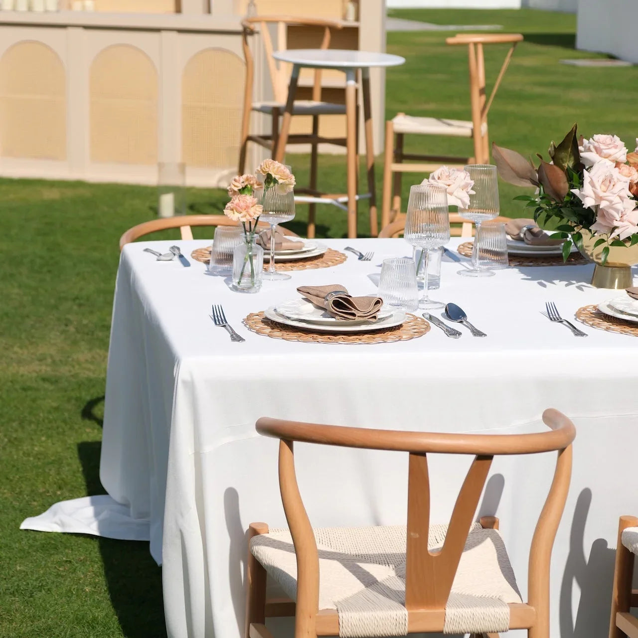 Luxe Alfresco - Dinner Setup featuring a white-clothed table for 8 with woven placemats, gold cutlery, glassware, floral centerpieces, and crossback chairs.