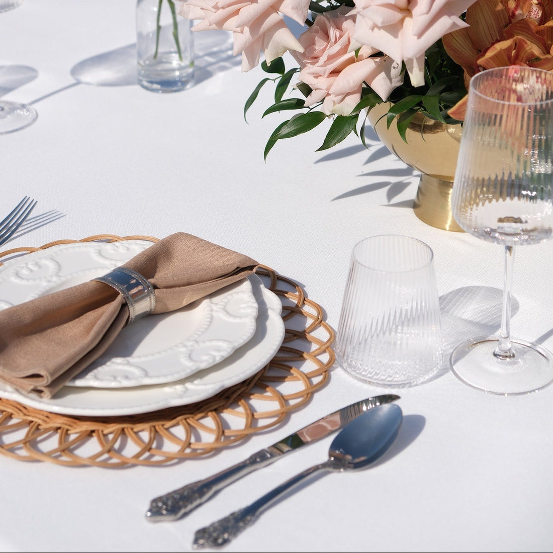 Luxe Alfresco - Dinner Setup featuring a table with rattan chargers, embossed china, gold cutlery, goblets, and blush rose centerpiece, perfect for elegant outdoor dining.