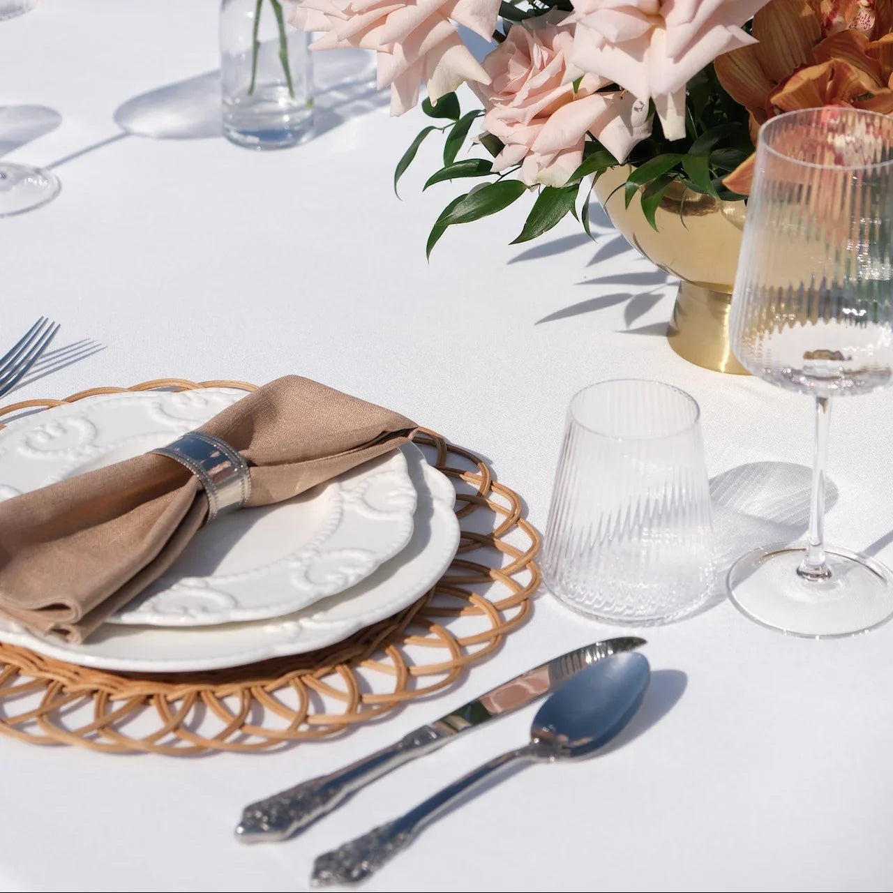 Luxe Alfresco - Dinner Setup featuring a table with rattan chargers, embossed china, gold cutlery, goblets, and blush rose centerpiece, perfect for elegant outdoor dining.