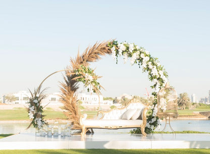 Round gold arch adorned with flowers, set behind a white couch with pillows, ideal for event decor.