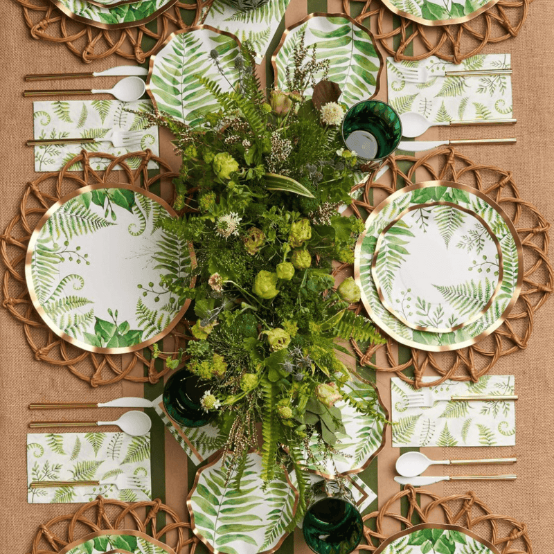 Disposable party tableware with green leaf plates, napkins, wooden cutlery, and woven placemats arranged around a greenery centrepiece.