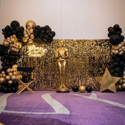 Golden Shimmer Celebration Wall with life-size statue, black and gold balloons, director's chair, star prop, and neon sign for Hollywood-themed events.
