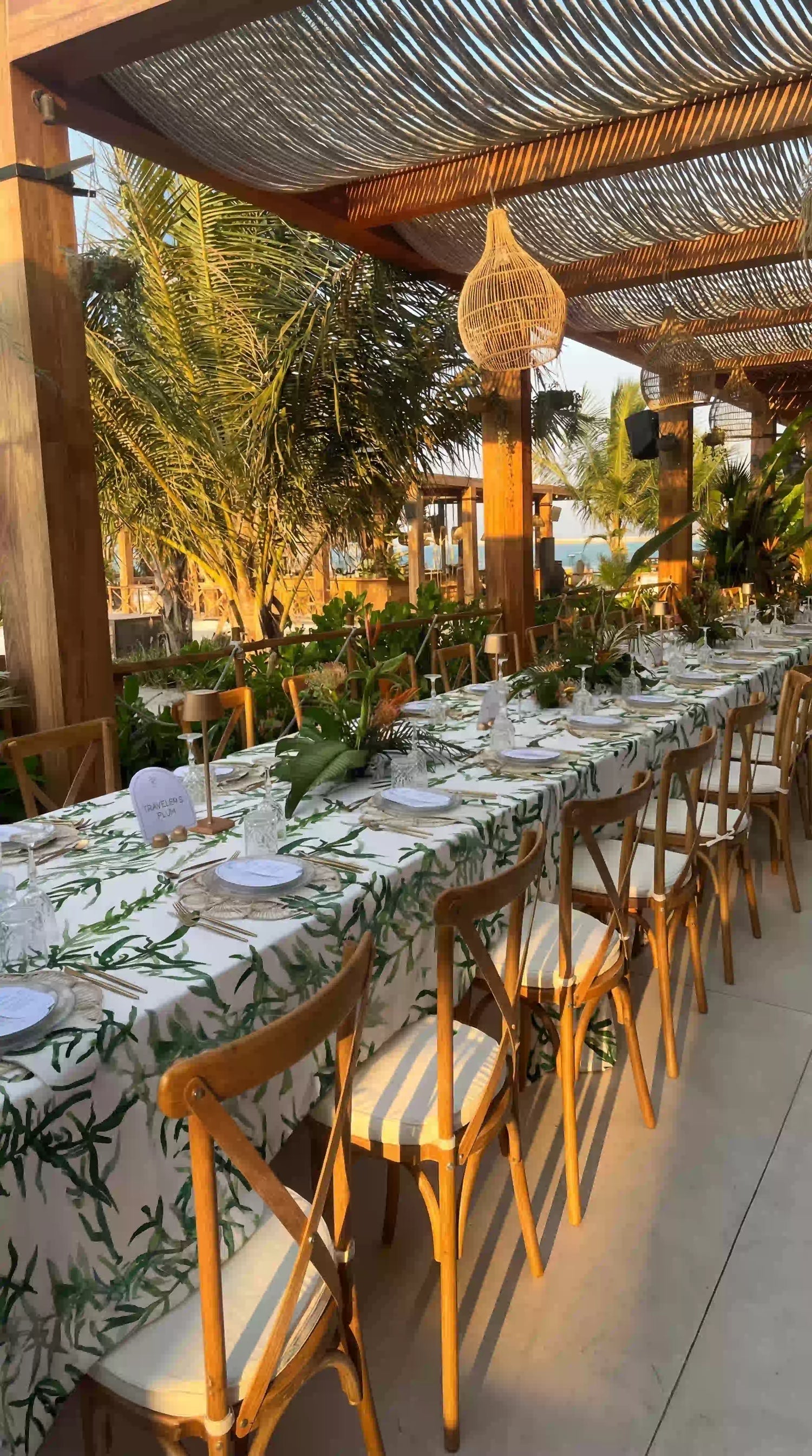 Gardenia Linen Tablecloth: Outdoor dining setup with a white tablecloth featuring green foliage, elegantly set with tableware under a pergola at a beachside event.