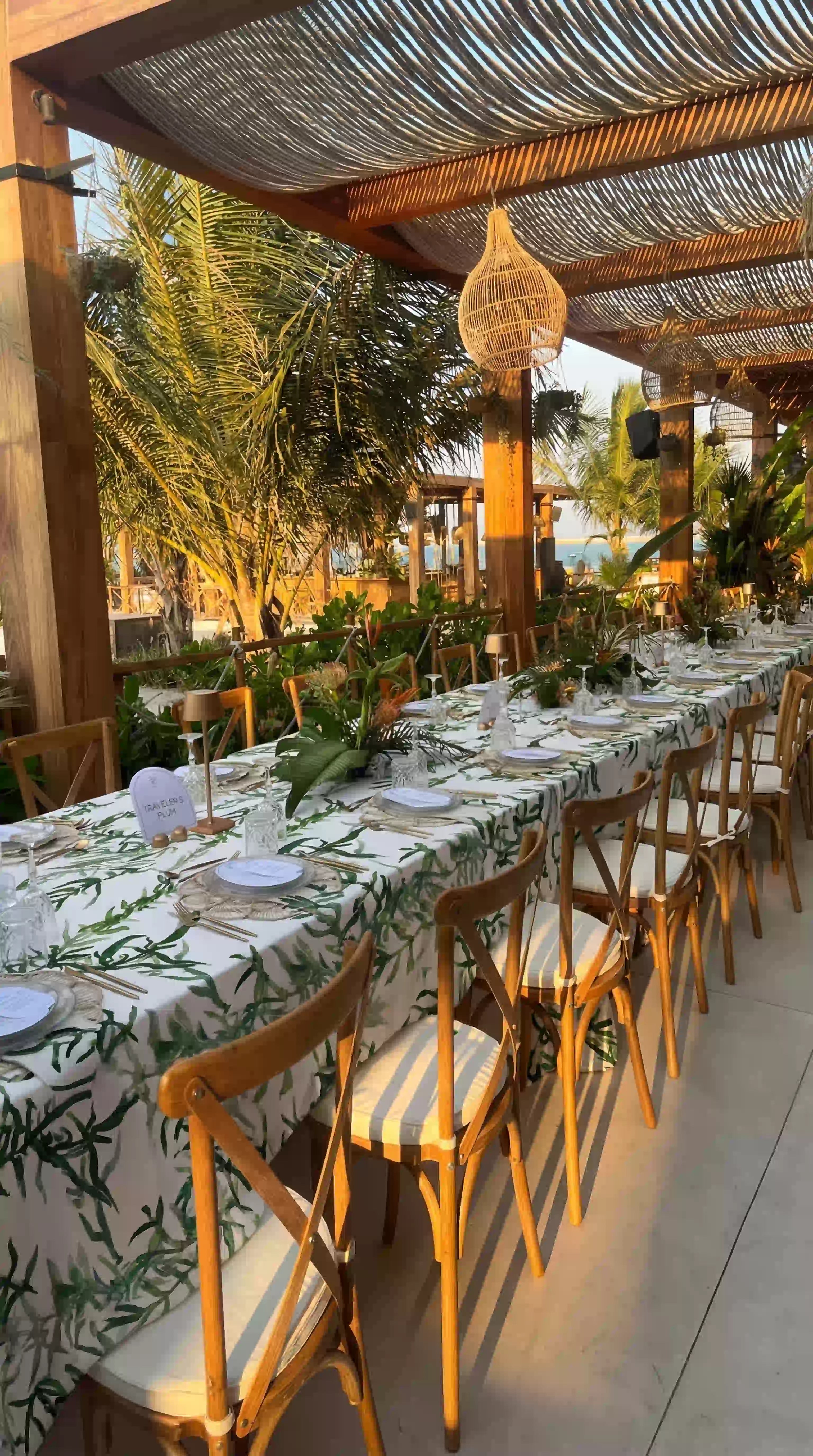 Gardenia Linen Tablecloth: Outdoor dining setup with a white tablecloth featuring green foliage, elegantly set with tableware under a pergola at a beachside event.