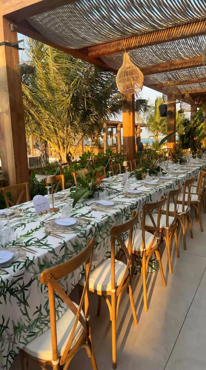 Gardenia Linen Tablecloth: Outdoor dining setup with a white tablecloth featuring green foliage, elegantly set with tableware under a pergola at a beachside event.