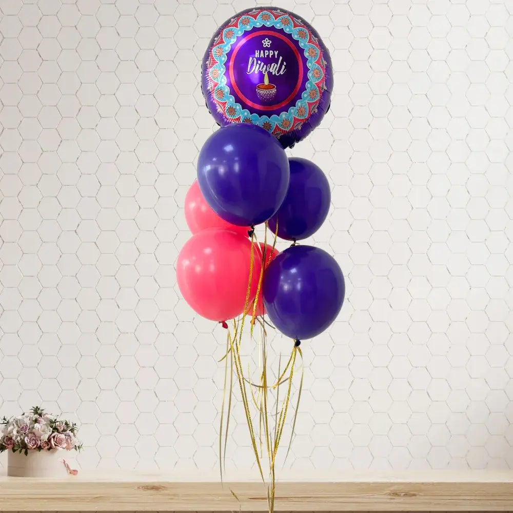 A festive balloon bouquet featuring a round metallic foil balloon with a Diwali design, surrounded by purple and pink latex balloons tied with gold ribbons against a white hexagonal wall.