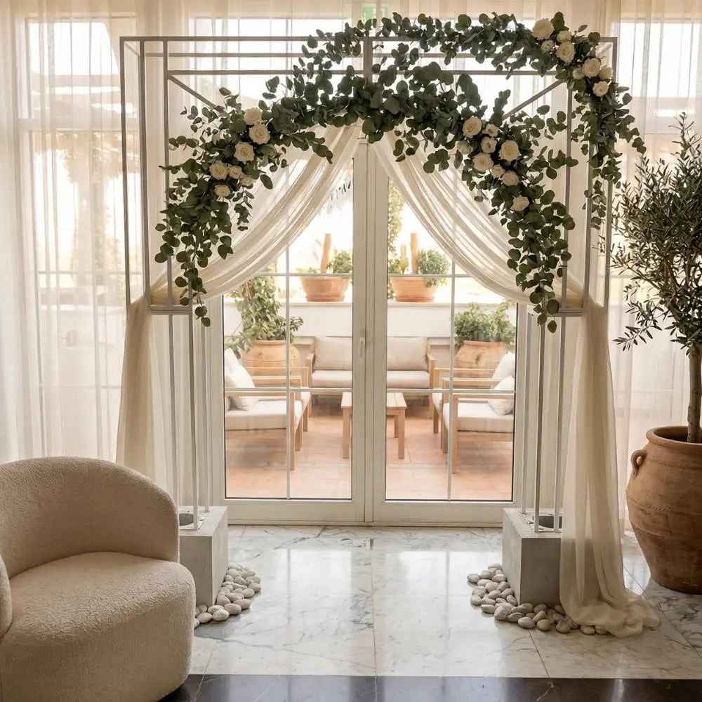 Geometric Metal Backdrop Arch draped in sheer fabric and eucalyptus, ideal for events, set against French-style glass doors with a cozy terrace view.